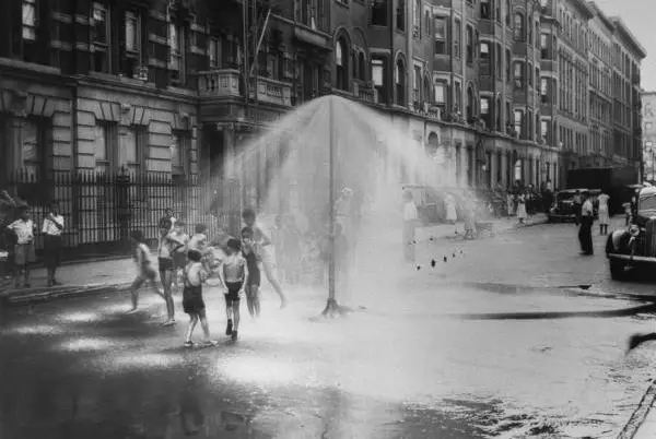 "Young children running around under sprinkler in street to cool off during summer, in South Harlem in shot from series of NYC Police Dept.'s Crime Prevention Photos of Juvenile Aid Bureau." August 8th, 1937.